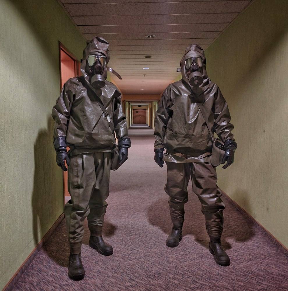 Two individuals wearing full olive-drab hazmat suits and gas masks stand in a long hotel hallway.